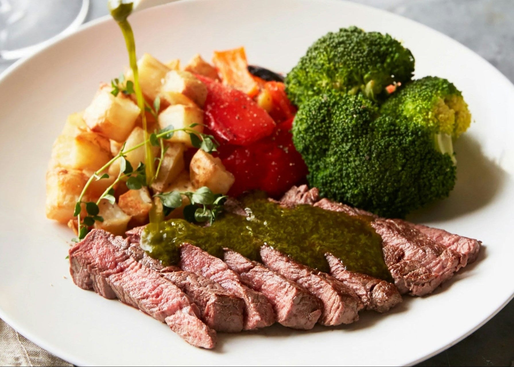 Person pouring green sauce over a plate of steak, vegetables, and pasta with a glass of red wine in the background.